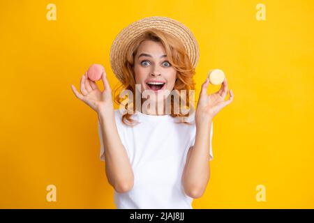 Nette junge Frau halten Makronen. Schönes lächelndes junges Mädchen, das bunte Makronen auf gelbem Hintergrund isst. Stockfoto