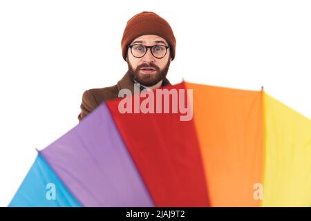 Studioporträt eines kaukasischen Hipster-Mannes in Herbstkleidung mit Mütze und Mantel, der seinen Regenbogenschirm mit einem überraschenden Gesichtsausdruck spinnt. Hochwertige Fotos Stockfoto