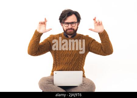 Ich hoffe, dass ich diese Tickets bekommen habe. Studio-Aufnahme eines kaukasischen bärtigen Millennials in einem Strickpullover mit einem Laptop auf den Knien, wobei die Daumen gedrückt wurden. Hochwertige Fotos Stockfoto