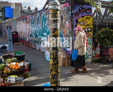 Ein Mann, der mit einem Kopftuch aus dem Nahen Osten bekleidet ist, kommt an einem Obststand am Brick Lane Market, London, England, Großbritannien vorbei Stockfoto