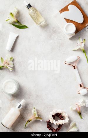 Flache Lay-Komposition aus natürlicher Hautpflegekosmetik, Gua Sha, Gesichtsmassagerolle, Blumen auf Steintisch. Stockfoto