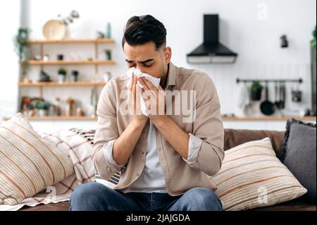 Unglücklicher, trauriger indischer oder arabischer Mann, der an Fieber und Grippe leidet, auf dem Sofa im Wohnzimmer sitzt, Nase bläst und in Serviette niest, eine Behandlung von Krankheiten braucht und Ärzte konsultieren muss Stockfoto