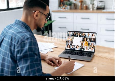 Fernunterricht, Online-Kurs. Blick über die Schulter eines Mannes, der sich in einem Notizbuch Notizen macht, auf einen Laptop-Bildschirm mit einem Lehrer und einer Gruppe von Studenten aus verschiedenen Rassen. E-Learning, Webinar, Online-Vortrag Stockfoto