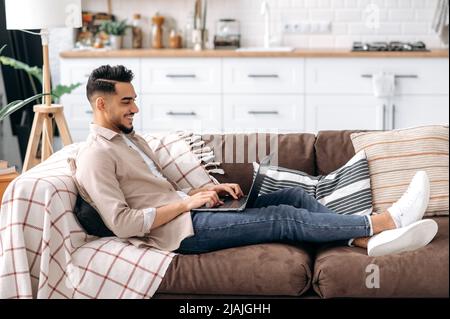 Schöner, moderner arabischer oder indischer Mann in lässiger, stilvoller Kleidung, Freiberufler oder Student, sitzend auf dem Sofa im Wohnzimmer, nutzt Laptop für die Arbeit an einem Projekt, elearning, Surfen im Internet, lächelnd Stockfoto
