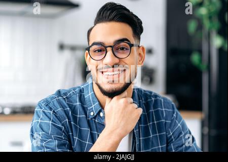 Nahaufnahme eines attraktiven, positiven arabischen oder indischen jungen Mannes mit Brille, Freiberufler oder Student, in legerer Kleidung, mit perfekten weißen Zähnen, der mit einem fröhlichen Lächeln auf die Kamera blickt Stockfoto