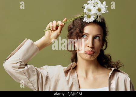 Lustige schöne verärgert lockig schöne Frau in Leinen casual Hemd halten chamomies Blumen über posiert isoliert auf über olivgrünen Pastellhintergrund Stockfoto