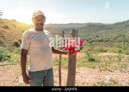 Mittellange Aufnahme eines reifen Latino-Mannes mit Schnurrbart und einer Mütze, die violette Blumen in den Händen hält, in einer Bergregion Nicaraguas Stockfoto