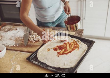 Eine Frau verteilt Tomatensauce auf eine Pizza, die zum Backen auf dem Ofen ausgelegt ist Stockfoto