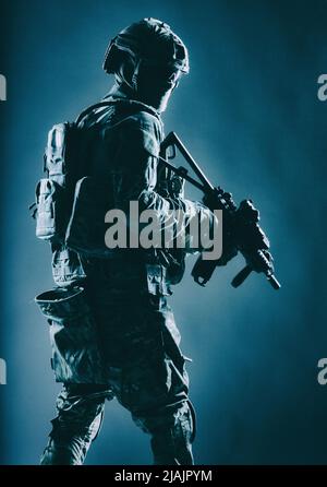 Armeesoldat in Kampfuniform mit Dienstgewehr in der Hand, blickt über die Schulter und schleicht sich in der Dunkelheit. Stockfoto