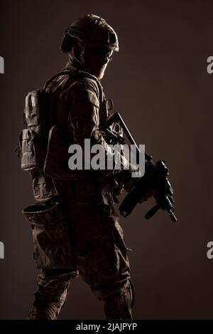 Armeesoldat in Kampfuniform mit Dienstgewehr in der Hand, blickt über die Schulter und schleicht sich in der Dunkelheit. Stockfoto