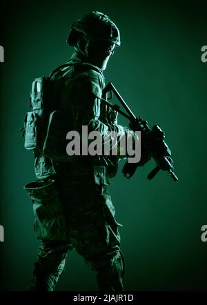 Armeesoldat in Kampfuniform mit Dienstgewehr in der Hand, blickt über die Schulter und schleicht sich in der Dunkelheit. Stockfoto