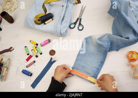 Kleidung ausbessern, Jean-Stoff mit Maßband messen, auf Arbeitstisch, Nähinstrumente wie farbige Fäden, Scheren, Handwerk Beruf Stockfoto