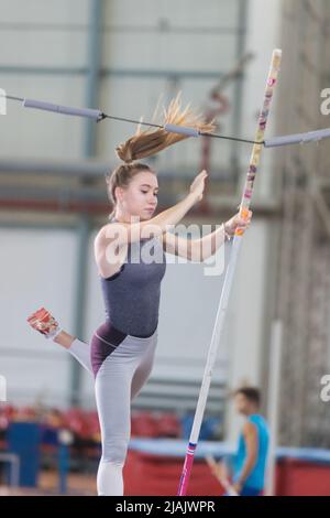 Stangenhochsprung im Innenbereich - junge sportliche Frau fällt nach dem Sprung über die Barriere herunter Stockfoto