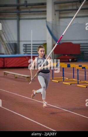 Stangenhochsprungungen im Innenbereich - junge sportliche Frau, die mit einem Stangenstock in den Händen auf der Startbahn läuft Stockfoto