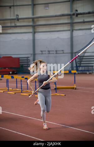 Stangenhochsprungungen im Innenbereich - junge sportliche Frau in Leggins, die mit einem Stock in den Händen läuft Stockfoto