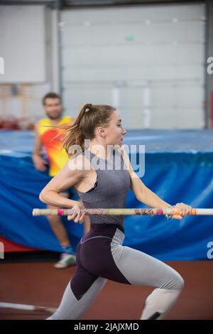 Stangenhochsprungungen im Innenbereich - junge sportliche Frau mit Pferdeschwanz, der mit einem Stock in den Händen läuft Stockfoto