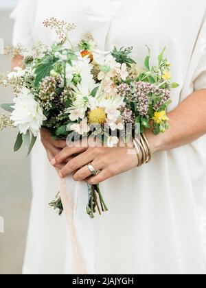Frau hält einen Hochzeitsstrauß mit einem Ring und Armbändern an. Strand Hintergrund mit einem minimalistischen Hochzeitskleid und Wildblumen in den Händen. Stockfoto