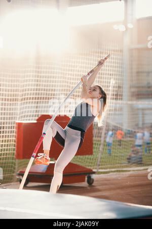 Stangenhochsprung im Innenbereich - junge sportliche Frau mit einem Pferdeschwanz, der zum Springen auf die Stange gelehnt ist - helles Licht auf dem Hintergrund Stockfoto