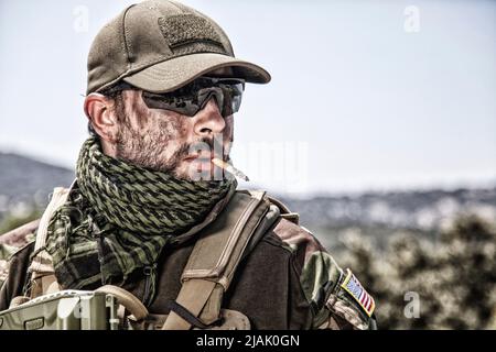 Soldat mit schmutzigem Gesicht raucht eine Zigarette, trägt ballistische Brille und Baseballmütze. Stockfoto