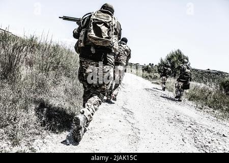 Soldaten, die eine Landstraße entlang laufen, zielen mit ihren Waffen auf Grasflächen. Stockfoto