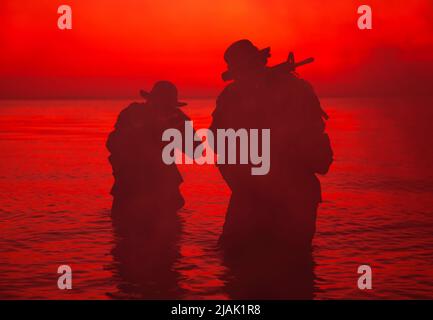 Silhouette von zwei Soldaten, die an der Küste watten und sich mit gezogenen Sturmgewehren in der Dunkelheit schleichen. Stockfoto