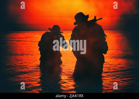 Silhouette von zwei Soldaten, die an der Küste watten und sich mit gezogenen Sturmgewehren in der Dunkelheit schleichen. Stockfoto