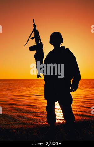 Silhouette eines Soldaten mit einem leichten Maschinengewehr an der Küste mit Sonnenuntergang Himmel. Stockfoto