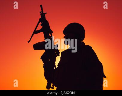 Silhouette eines Soldaten mit einem leichten Maschinengewehr bei Sonnenuntergang. Stockfoto