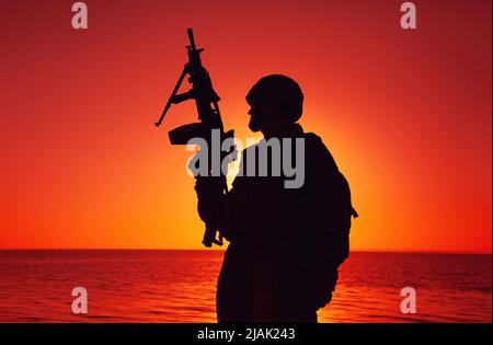 Silhouette eines Soldaten mit einem leichten Maschinengewehr an der Küste mit Sonnenuntergang Himmel. Stockfoto