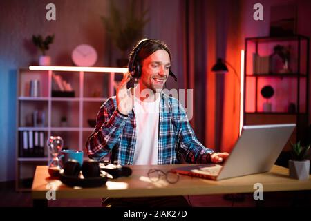 Junger Mann aus dem Kaukasus im Headset, der mit einem Laptop am Schreibtisch sitzt, Videoanruf führt oder einen Online-Schulungskurs führt. Geschäftsmann, der moderne Geräte für die Online-Sprachausgabe verwendet und online vom Heimbüro aus arbeitet Stockfoto