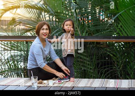 BANGKOK, THAILAND-FEBRUAR 23,2020:Unidentifizierte Großmutter kümmert sich um die Fütterung ihres Enkels Essen und Wasser, während faulenzen auf der Terrasse am Fluss in Hi Stockfoto