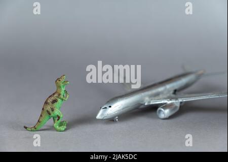 Kinderspielzeug. Silberne Flugzeug- und Dinosaurierminiatur vor grauem Hintergrund. Kleine grüne Figur eines räuberischen Dinosauriers, der auf seinem Hinterbein steht Stockfoto