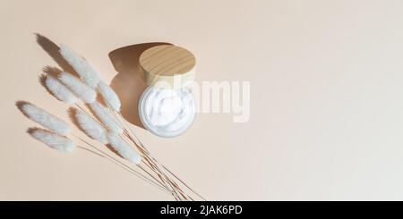 Feuchtigkeitscreme im offenen Glas und trockene Kräuter auf beigem Hintergrund, Draufsicht. Pflege empfindlicher Haut mit natürlichem Extrakt. Bunner mit Hautpflege bo Stockfoto