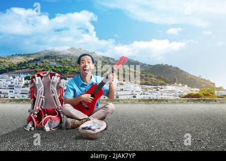 Asiatischer Mann mit Beanie-Hut und Rucksack, der Gitarre spielt, um auf der Straße mit blauem Himmel Geld zu verdienen Stockfoto