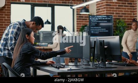 Gemischtes Team von Softwareentwicklern, die den Quellcode analysieren und auf Bildschirme zeigen, wobei der Algorithmus mit der Benutzeroberfläche auf einem digitalen Tablet verglichen wird. Programmierer, die am Codierungsgruppenprojekt arbeiten, sitzen am Schreibtisch. Stockfoto