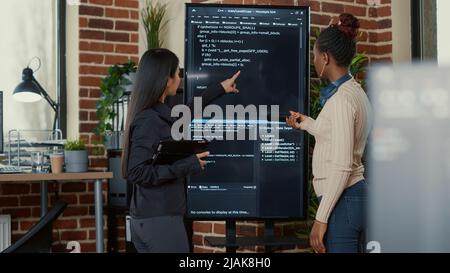 Team von Programmierern, die Code auf dem Wandbildschirm-tv analysieren, auf der Suche nach Fehlern und Fehlern, während sie digitales Tablet und Fernbedienung halten. Software-Ingenieure arbeiten an einem Gruppenprojekt für Cloud Computing zusammen. Stockfoto