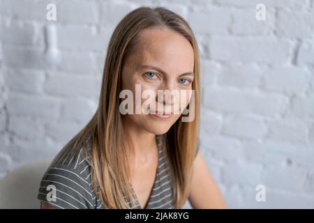 Porträt mit Schuss eine fröhliche blonde Frau mittleren Alters, die lächelt, wenn man die Kamera anschaut, fühlt sich in einer glücklichen Pose an, blauäugig in einem grauen Kleid an einer weißen Ziegelwand, Nahaufnahme Stockfoto
