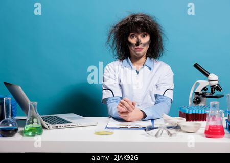 Verrückter verrückter Biochemie-Experte mit unordentlichen Haaren und schmutzigem Gesicht, der nach der Explosion des Experiments am Labortisch sitzt. Maniac goofy aussehende Chemiker dumm, während Blick auf die Kamera auf blauem Hintergrund. Stockfoto