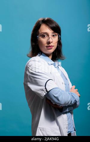 Biochemie-Spezialist trägt Schutzbrille und Laborkittel, der auf blauem Hintergrund steht, während er auf die Kamera schaut. Biomedizinischer Forscher mit gekreuzten Armen auf blauem Hintergrund, Aufnahme im Studio. Stockfoto