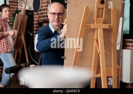 Porträt eines älteren Mannes, der in der Draw-Klasse vor auf Leinwand sitzt, Zeichnung Vasenmodell mit Bleistift. Vielfältiges Team, das an der Skizzierungstechnik arbeitet und künstlerische Fähigkeiten entwickelt. Kreatives Konzept Stockfoto
