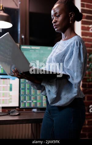 afroamerikanischer Devisenmarkthandelsagent mit einem Laptop, der vor einer Multi-Monitor-Workstation steht. Maklerfirma Mitarbeiter mit Computer während der Analyse von Live-Grafiken Stockfoto