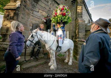 Der zeremonielle König Jon Haddock wird dabei unterstützt, während der Castleton Garland eine große Girlande abzuheben, eine alte Tradition, bei der der König einen großen, mit Blumen bedeckten Rahmen anzieht, bevor er eine Prozession zu Pferd durch das Dorf Castleton im Peak District National Park in Derbyshire führt. Bilddatum: Montag, 30. Mai 2022. Stockfoto