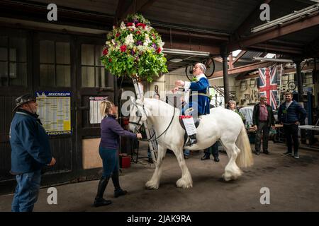 Der zeremonielle König, Jon Haddock, wird während der Castleton Garland in eine große Girlande verholfen, eine alte Tradition, bei der der König einen großen, mit Blumen bedeckten Rahmen anzieht, bevor er eine Prozession zu Pferd durch das Dorf Castleton im Peak District National Park in Derbyshire führt. Bilddatum: Montag, 30. Mai 2022. Stockfoto