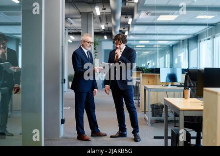 Reifer Geschäftsmann im Anzug, der seinem Kollegen etwas auf einem digitalen Tablet zeigt, während er im Büro steht Stockfoto
