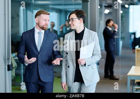 Zwei junge Kollegen sprechen miteinander, während sie durch das moderne Büro gehen Stockfoto