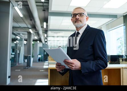 Porträt eines älteren Geschäftsmannes in dunklem Anzug, der Bericht hält und die Kamera anschaut, während er im Bürogebäude steht Stockfoto