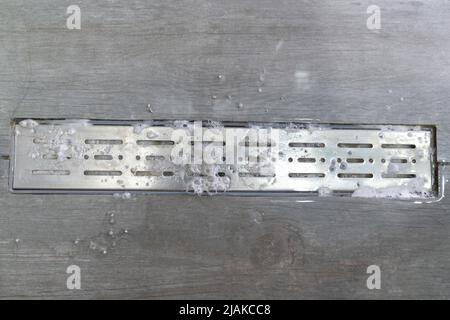 Ablaufloch mit Rohrreiniger im Metallbecken. Schaum mit Seifenblasen auf dem Boden in der Dusche Stockfoto