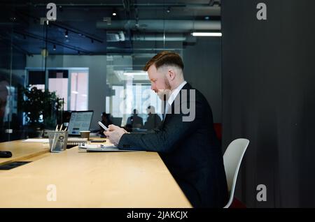 Junger Geschäftsmann in formellem Anzug, der im Sitzungssaal am Tisch sitzt und mit dem Mobiltelefon auf die Besprechung wartet Stockfoto