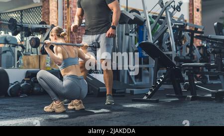 Eine hübsche junge Frau aus europa, die eine Langhantel auf ihren Schultern hält und knirscht. Nicht erkennbarer Mann in T-Shirt und Shorts, der neben ihr steht und jubelt. Hochwertige Fotos Stockfoto