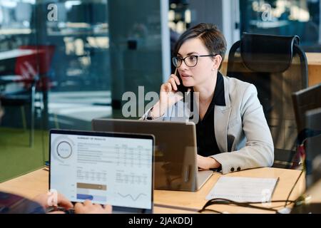 Junge Geschäftsfrau in Brillen, die mit einem Laptop am Arbeitsplatz ein Gespräch auf dem Mobiltelefon führt Stockfoto